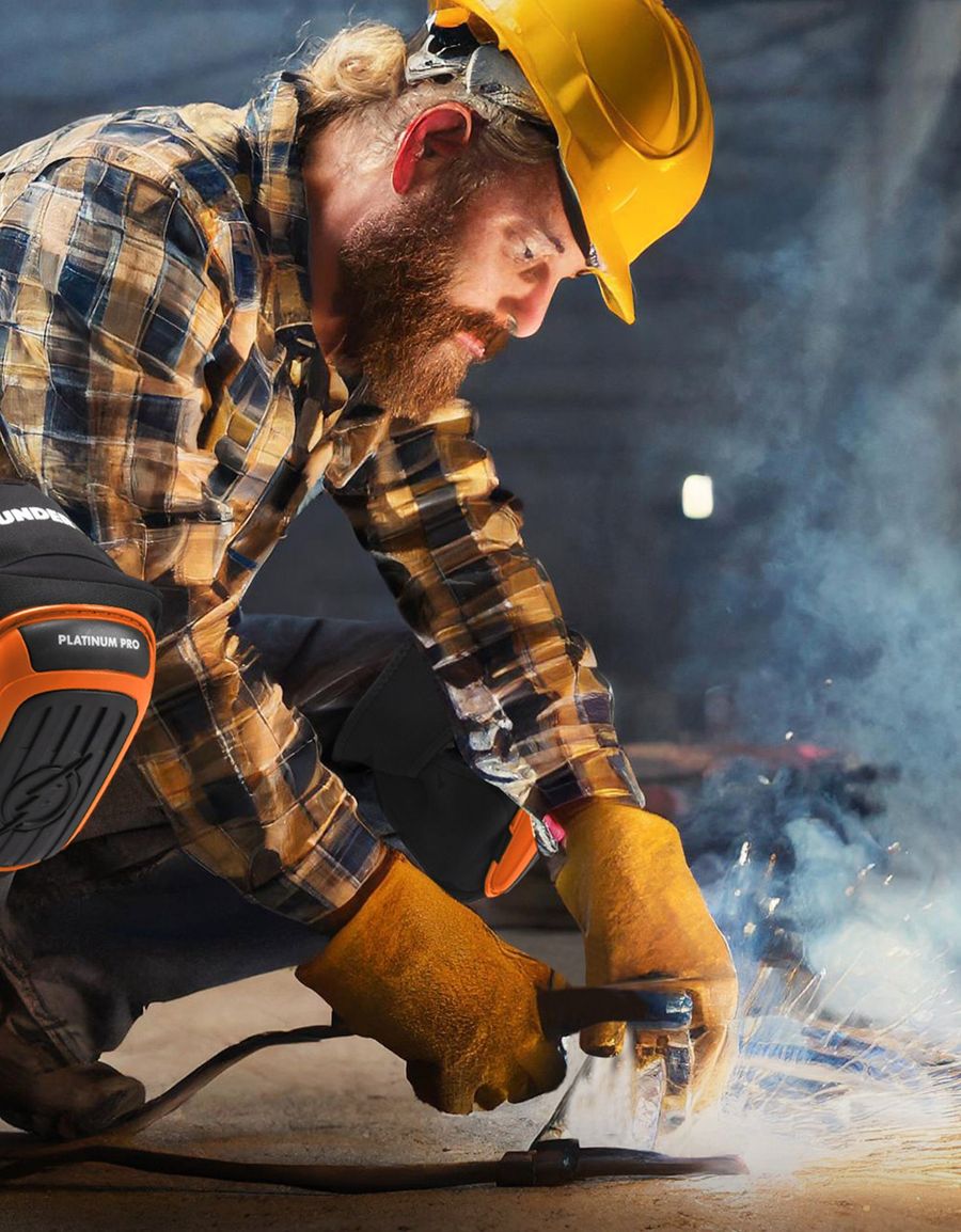 Welder in a hard hat kneeling on a concrete shop floor in orange Thunderbolt Platinum Pro knee pads